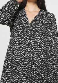 Musta mekko V-pääntiöllä, jossa on valkoinen abstrakti raitakuvio. Pitkät puffi hihat ja virtaava siluetti.