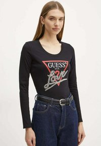 Camisa de manga larga negra con un gráfico triangular rojo que dice "GUESS U.S.A." y detalles plateados decorativos, combinada con jeans de mezclilla azules y un cinturón negro.