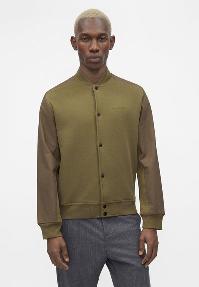 Άνδρας με κοντά ξανθά μαλλιά που φοράει μπουφάν bomber σε λαδί και καφέ χρώμα με κουμπιά και γκρι παντελόνι, στέκεται σε λευκό φόντο.