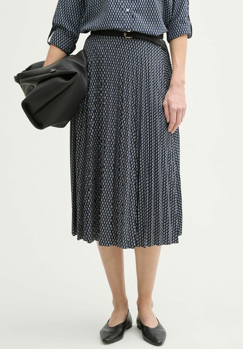 Femme portant une jupe plissée à motifs et une chemise assortie, taille marquée par une ceinture, tenant un grand sac en cuir noir plié, portant des ballerines noires.