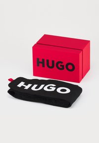 Juodas galvos apdangalas su užrašu "HUGO" baltomis raidėmis, gulintis šalia raudonos stačiakampės dėžutės su "HUGO" drąsiomis juodomis raidėmis.