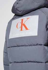 Szara kurtka puchowa z kapturem, z dużą białą metką z czerwonym napisem "CK" oraz tekstem "Calvin Klein Jeans" na plecach.