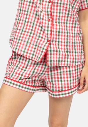 Rood, groen en wit geruit button-up pyjamatopje en bijpassende short met rode bies, gedragen door een persoon die voor een effen achtergrond staat.