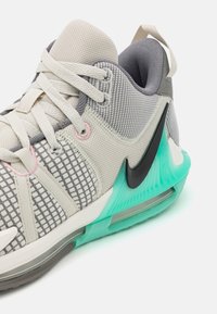 Svetle šedá sportovní bota s průsvitným svrškem, mřížkovaným vzorem a tyrkysovými akcenty. Obsahuje černé logo swoosh a polstrovanou podrážku pro podporu.