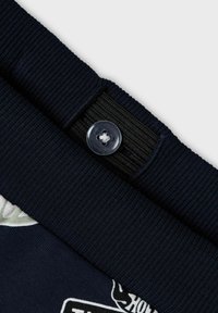 Tessuto blu navy con texture a coste, caratterizzato da un evidente bottone trasparente a quattro fori e una fascia elastica nera. La superficie presenta un motivo grafico in colori chiari.