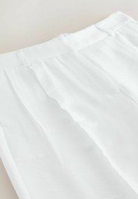 Pantalon blanc taille haute avec passants de ceinture, plis et fermeture éclair à l'avant sur un fond neutre.