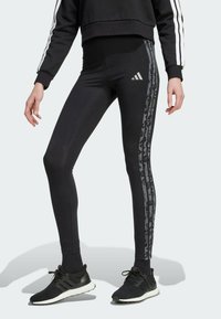 Black leggings with a high waist and a slim fit, featuring dark camo stripes down the sides and an Adidas logo in silver.