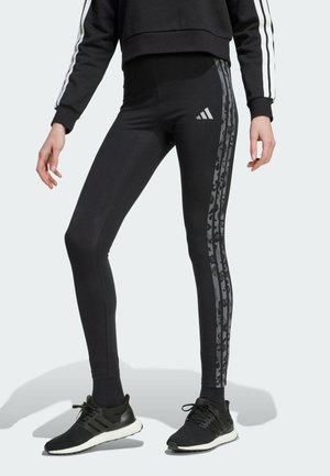 Leggings noirs avec taille haute et coupe slim, présentant des rayures camouflées sombres sur les côtés et un logo Adidas en argent.