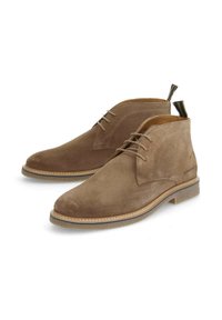 Bottines en daim marron avec un bout arrondi, un laçage fin et une semelle contrastante. Elles comportent une languette en boucle à l'arrière.