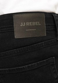Jeans in denim nero con un'etichetta in pelle con "JJ REBEL" in rilievo. La vita ha una finitura pulita con cuciture visibili e una texture sottile.