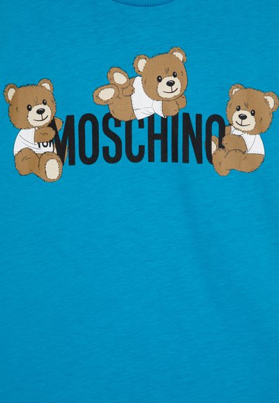 Camiseta azul con tres osos de dibujos animados que llevan camisetas blancas, acompañados del texto "Moschino" en una fuente negra en negrita. Tejido suave y texturizado.