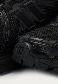 Gros plan sur des chaussures de sport noires en mesh texturé et cuir, avec des semelles épaisses et un logo "N" en relief sur le côté et la semelle.
