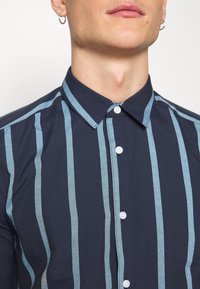 Camicia blu navy con strisce verticali azzurre chiare, colletto con bottoni e bottoni bianchi. Tessuto liscio con vestibilità su misura.
