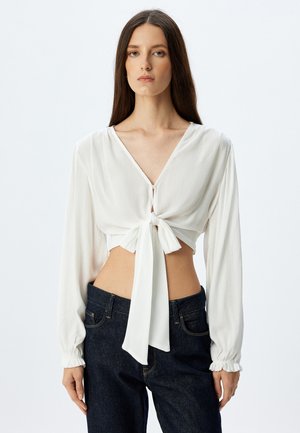 Blusa bianca cropped con scollatura a V profonda, annodata davanti con un fiocco. Maniche lunghe con polsini arricciati, realizzata in tessuto leggero e testurizzato.