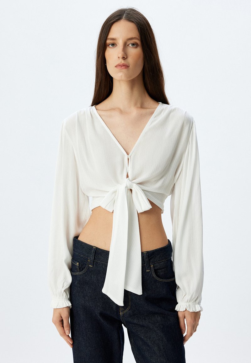 Blusa bianca cropped con scollatura a V profonda, annodata davanti con un fiocco. Maniche lunghe con polsini arricciati, realizzata in tessuto leggero e testurizzato.