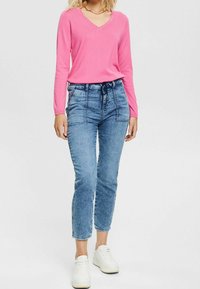 Różowa bluzka z długim rękawem i dekoltem w serek, zestawiona z wysokim stanem jasnoniebieskich dżinsów z paskiem wiązanym na sznurek oraz dopasowanymi nogawkami do kostki. Białe trampki.