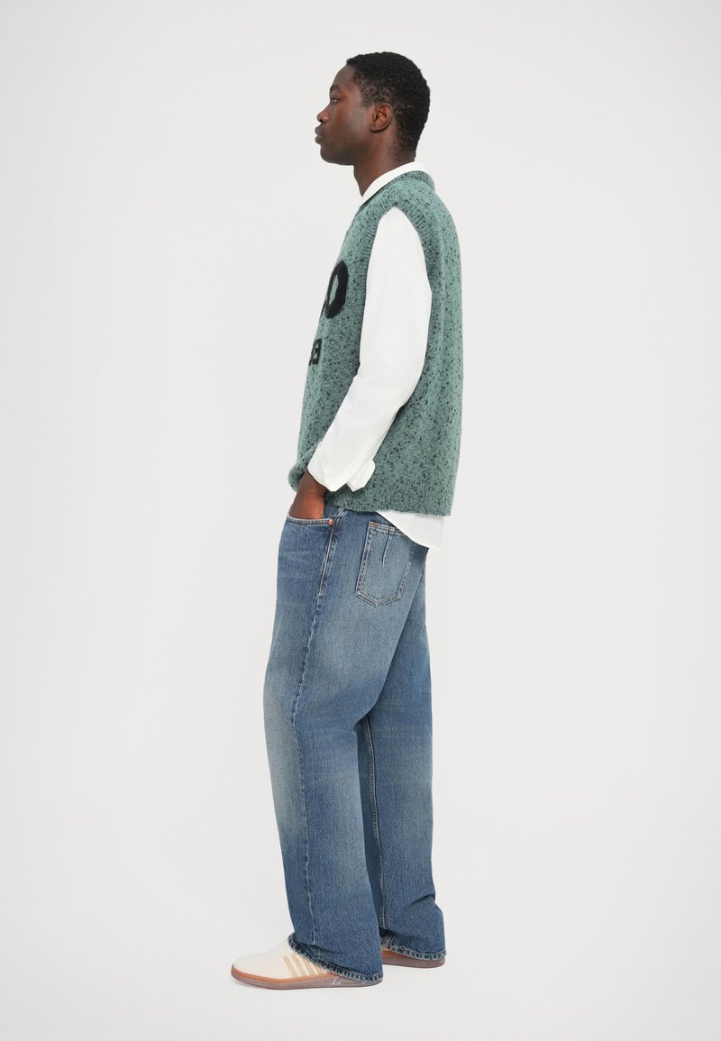 Gilet lavorato a maglia verde chiaro con testo nero, indossato sopra una maglietta a maniche lunghe bianca, abbinato a jeans in denim blu larghi e mocassini a righe.