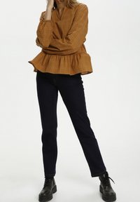 Camicia peplum a quadretti marrone con maniche lunghe e scollo a V, abbinata a jeans blu scuro e stivali neri con lacci.