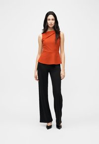 Kvinne som står iført ermeløs, rust-rød peplum-topp, svarte bukser med vide ben og svarte spisse hæler mot hvit bakgrunn.