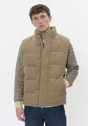 Tan corduroy puffer vest with high collar, snap closure, and two side pockets. Worn over a striped sweater, with blue denim jeans.