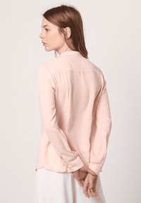 Camisa de manga larga color rosa pálido con cuello, textura suave y detalles fruncidos en los puños; dobladillo ligeramente curvado.