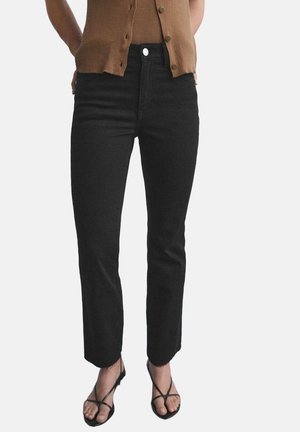 Vrouw draagt zwarte rechte jeans, bruin mouwloos topje met knopen en zwarte sandalen met bandjes, staand met handen achter de rug.