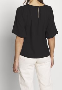 Černá blůza s nabíranými rukávy, strukturovaná tkanina, zapínání na klíčovou dírku na zádech a kulatý výstřih; kombinována se světle zbarvenými kalhotami.