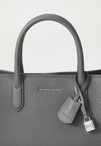 Bolso de cuero gris con textura de guijarros y dos asas en la parte superior, que presenta un charm plateado con el logo de MK y suturas sutiles en la parte frontal.
