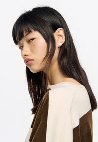 Model w beżowo-brązowej bluzce z geometrycznymi blokami kolorów. Małe okrągłe kolczyki i proste, ciemne włosy z grzywką.