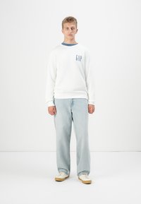 Joven de pie vistiendo un sudadera blanca de GAP, jeans azul claro y zapatillas blancas frente a un fondo claro.