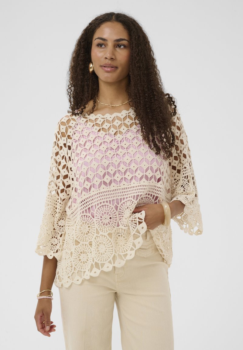 Femme portant un haut en dentelle crochetée beige sur un t-shirt rose avec un pantalon beige, accessoirisée avec des boucles d'oreilles créoles dorées, un bracelet et un collier.