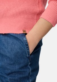 Rosa gerippter Strickpullover mit Dreiviertelärmeln, kombiniert mit blauen Jeans. Etikett nahe dem Saum sichtbar, Hand in der Tasche.