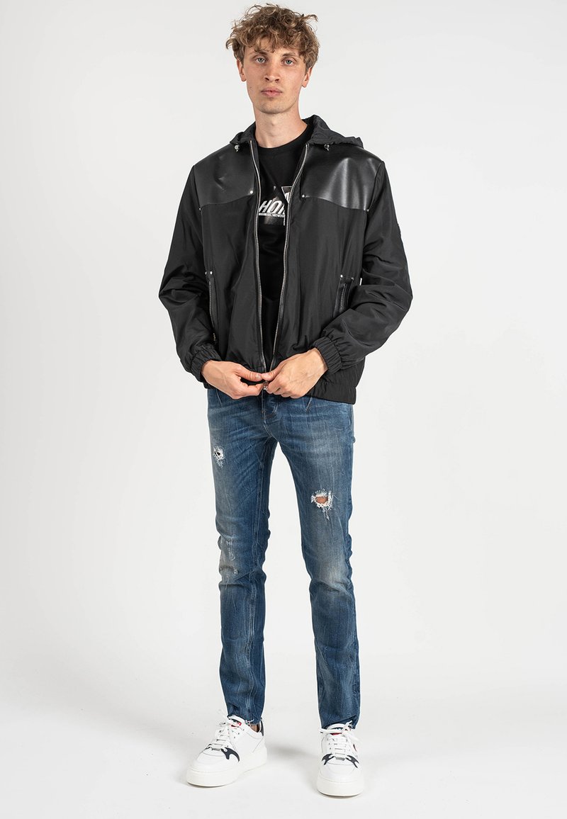 Giacca bomber nera con pannelli lucidi sul petto, polsini elasticizzati e due tasche con cerniera, abbinata a jeans blu strappati e sneakers bianche.