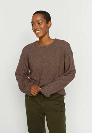 Personne souriante aux cheveux courts portant un pull marron en matière duveteuse et un pantalon vert olive sur un fond clair uni.
