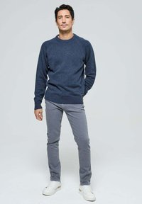 Pull marin avec une texture mouchetée, associé à un pantalon slim gris clair et des baskets blanches, se tenant devant un fond neutre.
