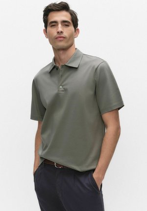 Man som bär en olivgrön kortärmad pikétröja instoppad i mörkblå byxor med händerna i fickorna, stående mot en enkel bakgrund.