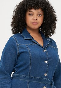 Casaco de ganga com gola, fecho de botão e cintura ajustada. O tecido é de um azul médio com uma textura subtil.