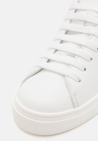 Sneaker bianco con tomaia in pelle liscia, punta tonda, lacci bianchi piatti e suola in gomma testurizzata. Design minimalista senza loghi visibili.