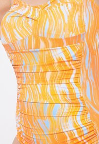 Zwemkleding in oranje met blauwe en witte golvende patronen. Kenmerkt zich door een gegatherde textuur, zachte stof en een ontwerp met één schouder.