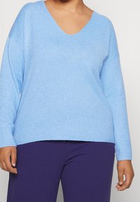 Lichtblauwe oversized trui met een V-hals, ribbelmanchetten en zoom, gemaakt van zacht materiaal. Gecombineerd met donkerpaarse broek.