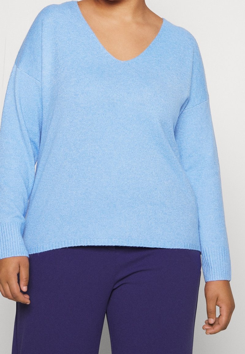 Jasnoniebieski oversize sweter z dekoltem w kształcie V, z prążkowanymi mankietami i dołem, wykonany z miękkiego materiału. Połączony z ciemnofioletowymi spodniami.