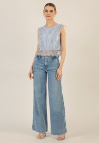 Helles blaues Spitzentop mit gewellten Ärmeln über hochgeschnittenen, weiten Denim-Jeans. Das Model steht in Riemchensandalen vor einem neutralen Hintergrund.