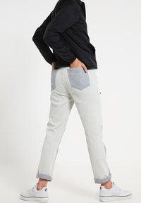 Jasnoszare dżinsy z kontrastowymi niebieskimi kieszeniami, podwiniętymi nogawkami i luźnym krojem. Noszone z czarną kurtką i białymi trampkami.