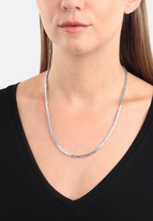 Primo piano di una donna che indossa una collana di catena argentata sopra una maglietta nera a V, con i capelli castani lisci visibili su un lato.