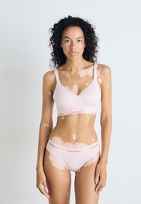 Világos rózsaszín Calvin Klein melltartó és bugyi szett vékony pántokkal, logóval ellátott rugalmas pánttal és sima textúrával. Modell egy sima háttér előtt áll.