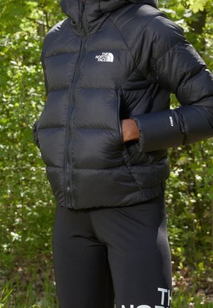 Osoba w czarnej kurtce puchowej The North Face i legginsach, stojąca na zewnątrz z rękami w kieszeniach kurtki.