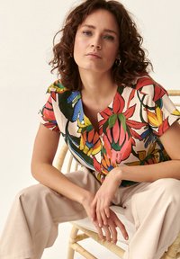 Top cu mânecă scurtă florală, cu culori vibrante precum roșu, galben și albastru pe un fundal crem, cu închidere cu butoni și guler rotund.