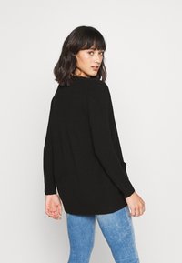 Svart strikkegenser med lange ermer, en løs passform og sidelommer. Båret over lyseblå jeans som har en teksturert finish.