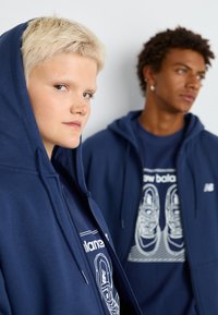 Suaras azuis marinhos com capuz usadas sobre t-shirts gráficas em cores contrastantes; uma das camisetas apresenta uma ilustração de um tênis. Tecido texturizado visível.