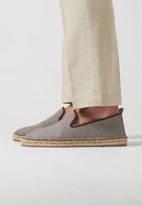 Alpargatas de tela con un patrón de cuadros en blanco y negro, acentos en marrón y suela de yute trenzado. Diseño suave y fácil de poner.